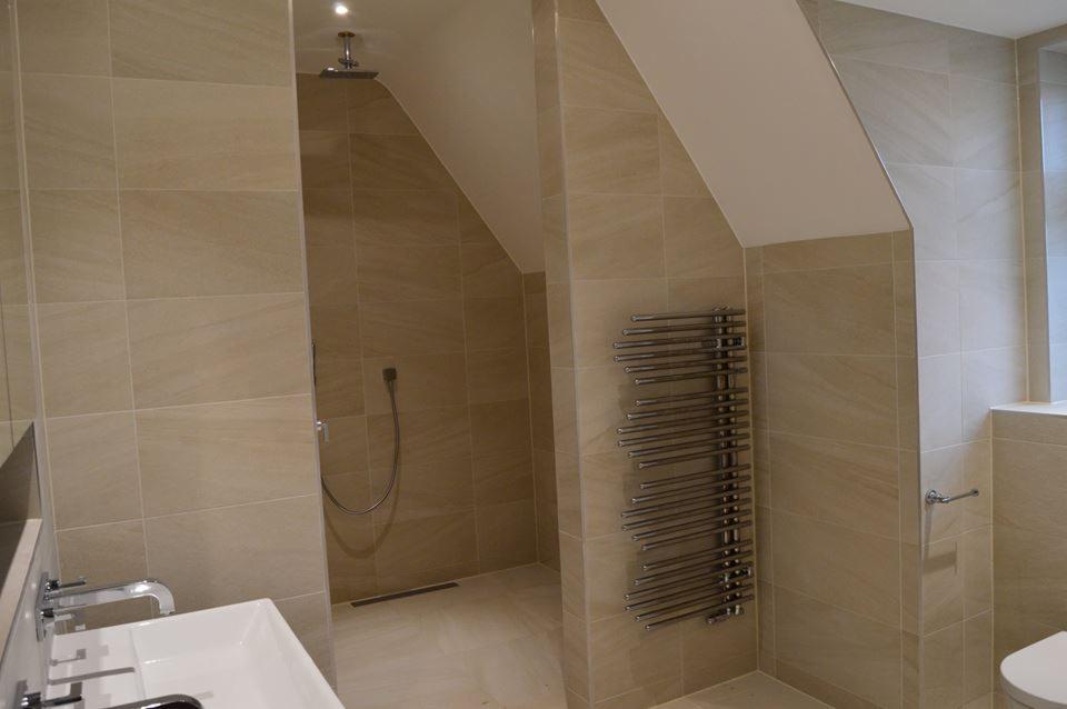 Łazienka na poddaszu z beżowymi płytkami, kabina prysznicowa typu walk-in z odpływem liniowym, grzejnik drabinkowy i umywalka z chromowaną armaturą.