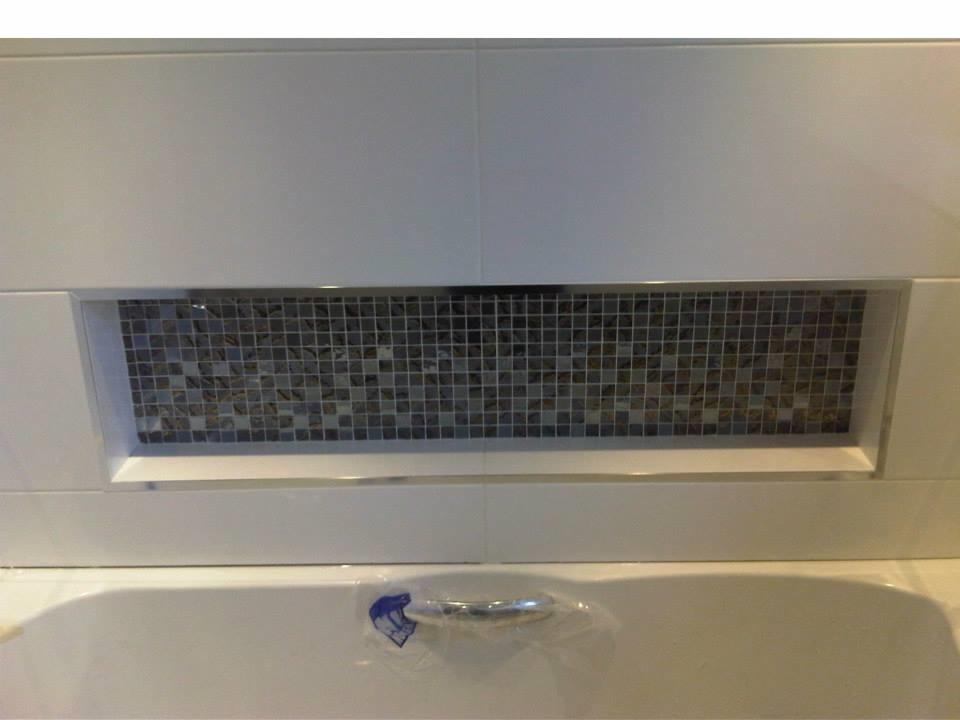 Białe, błyszczące płytki ścienne z wnęką wypełnioną mozaiką z drobnych, kwadratowych płytek w odcieniach szarości i brązu, nad białą umywalką z widocznym fragmentem baterii i naklejką zabezpieczającą.