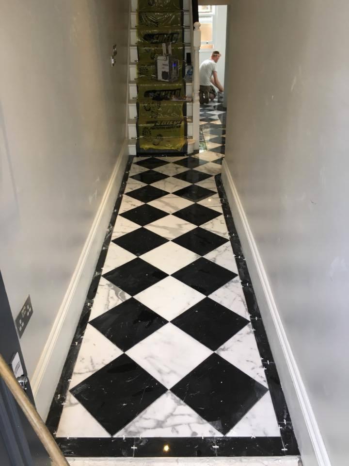 Korytarz z ułożoną podłogą w czarno-białą szachownicę z marmuru; na schodach folia ochronna, w tle widoczna osoba pracująca przy podłodze, widoczne krzyżyki dystansowe między płytkami.