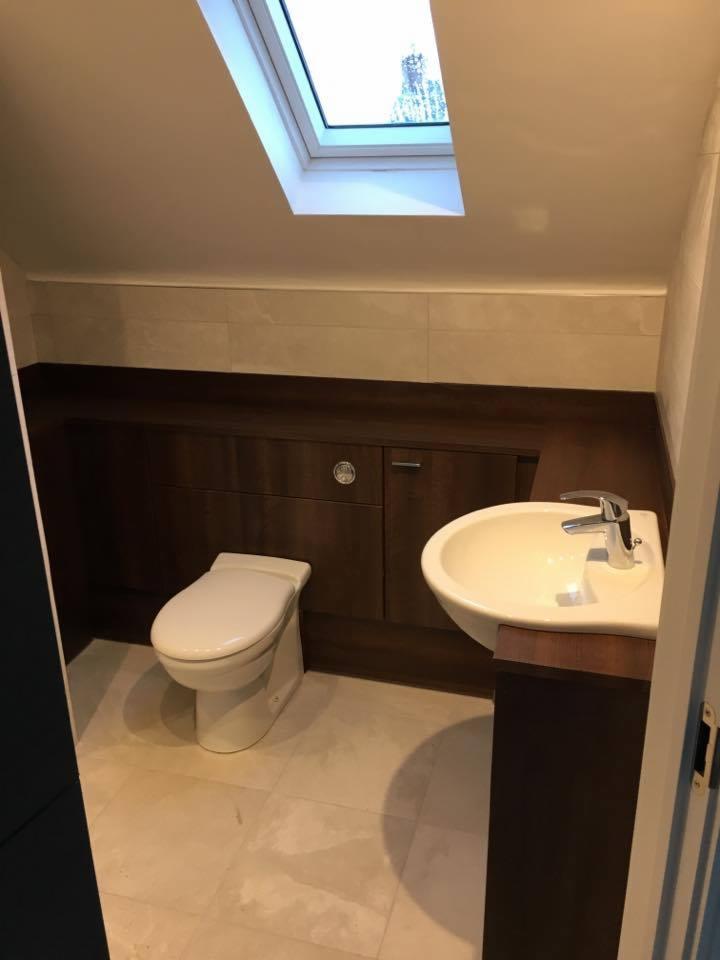 Nowoczesna łazienka na poddaszu ze skośnym sufitem i oknem dachowym, kremowe płytki podłogowe i ścienne, zabudowa z ciemnego drewna z umywalką i toaletą, chromowana bateria.
