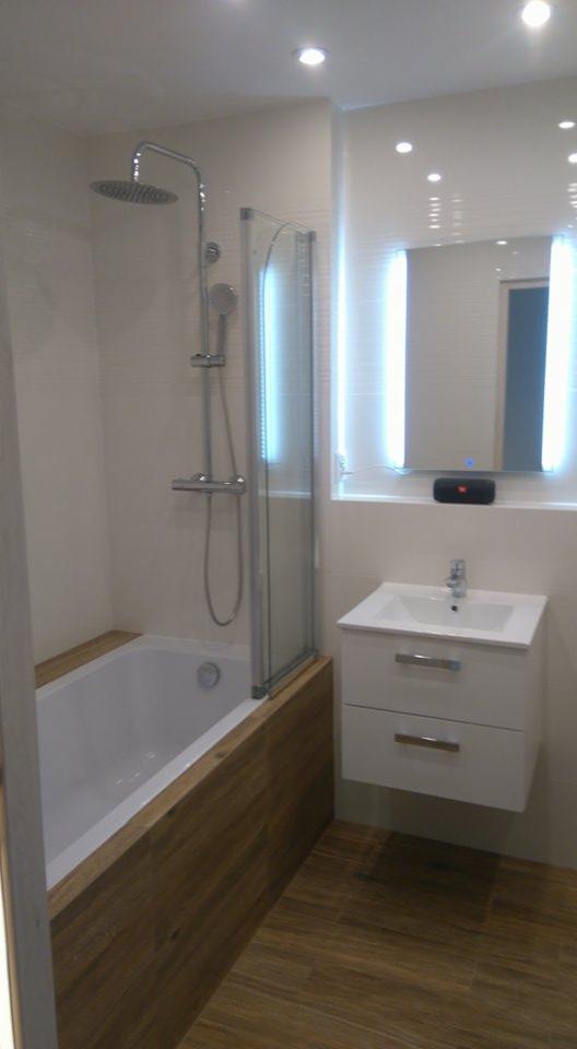 Nowoczesna łazienka z wanną obudowaną drewnopodobnymi panelami, szklanym parawanem nawannowym i podświetlanym lustrem nad białą szafką z umywalką. Na ścianie widoczny głośnik bezprzewodowy.