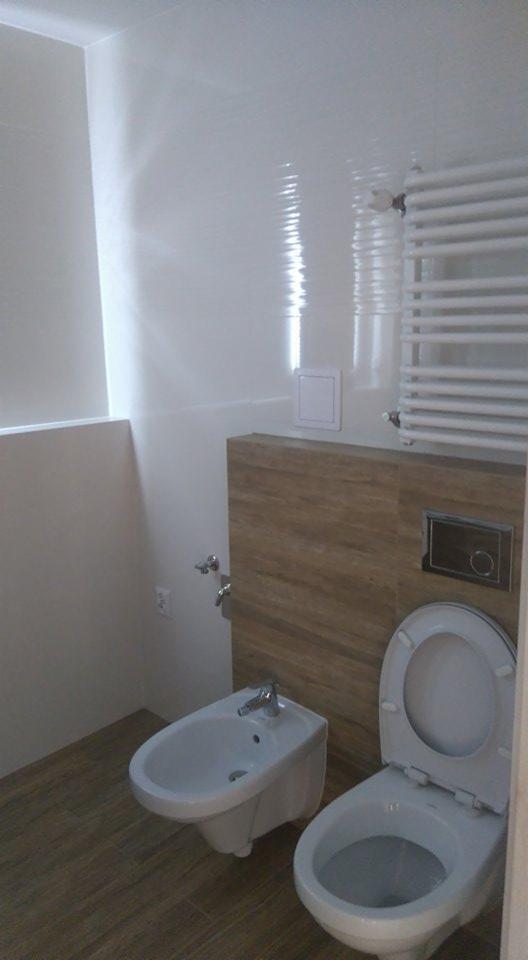 Nowoczesna łazienka po remoncie z białymi, błyszczącymi płytkami na ścianach, drewnopodobną zabudową, wiszącą toaletą i bidetem.