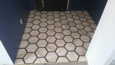 Podłoga wyłożona heksagonalnymi płytkami w odcieniach beżu z ciemnymi fugami, widok z góry na fragment pomieszczenia z granatową ścianą i białą kolumną.