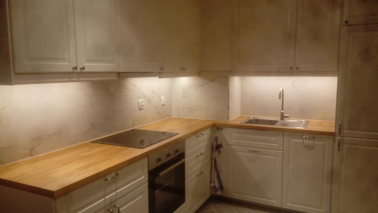 Jasna kuchnia z drewnianym blatem i marmurowym backsplashiem, oświetlona od dołu szafek górnych, z widoczną płytą indukcyjną i piekarnikiem.