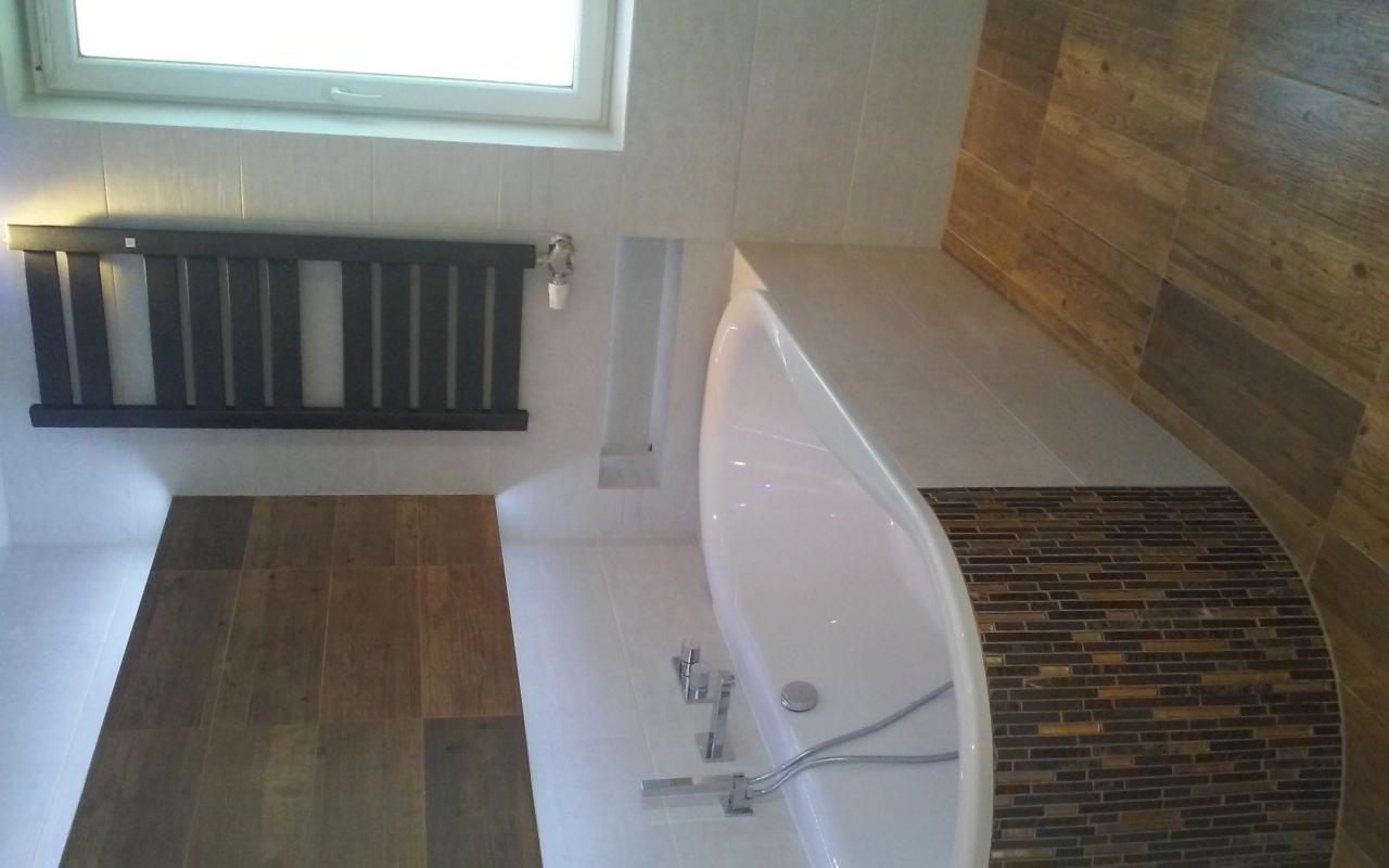 Biała wanna wolnostojąca z mozaiką w odcieniach brązu, drewnopodobne płytki na ścianie, grzejnik drabinkowy w łazience z drewnianą podłogą.