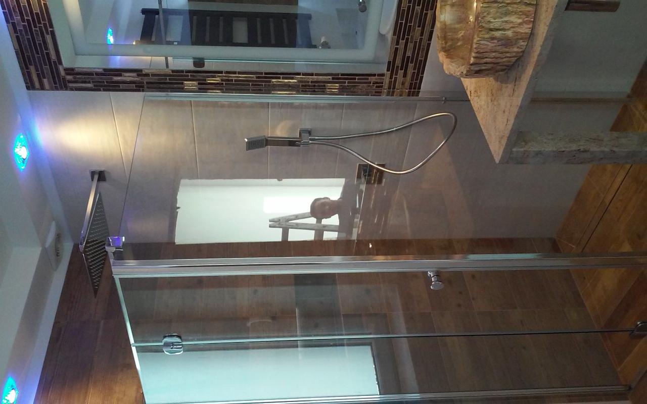 Nowoczesna kabina prysznicowa z deszczownicą i baterią, wykończona płytkami w odcieniach brązu i beżu, z elementami dekoracyjnymi z mozaiki i podświetleniem LED, widoczne odbicie osoby na drabinie...