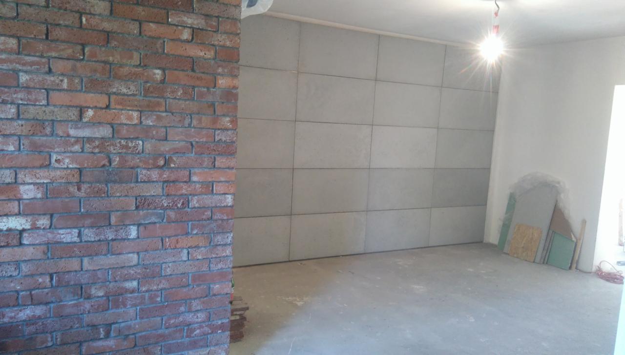 Wnętrze w trakcie prac remontowych: ściana z cegły klinkierowej, szare płytki ścienne ułożone w regularny wzór, surowy beton na podłodze, widoczne materiały budowlane w rogu pomieszczenia.