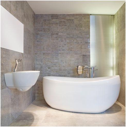 Nowoczesna łazienka z owalną wanną, wiszącą umywalką i szarą okładziną ścienną imitującą kamień, oświetlona przez pionowe żaluzje okienne.