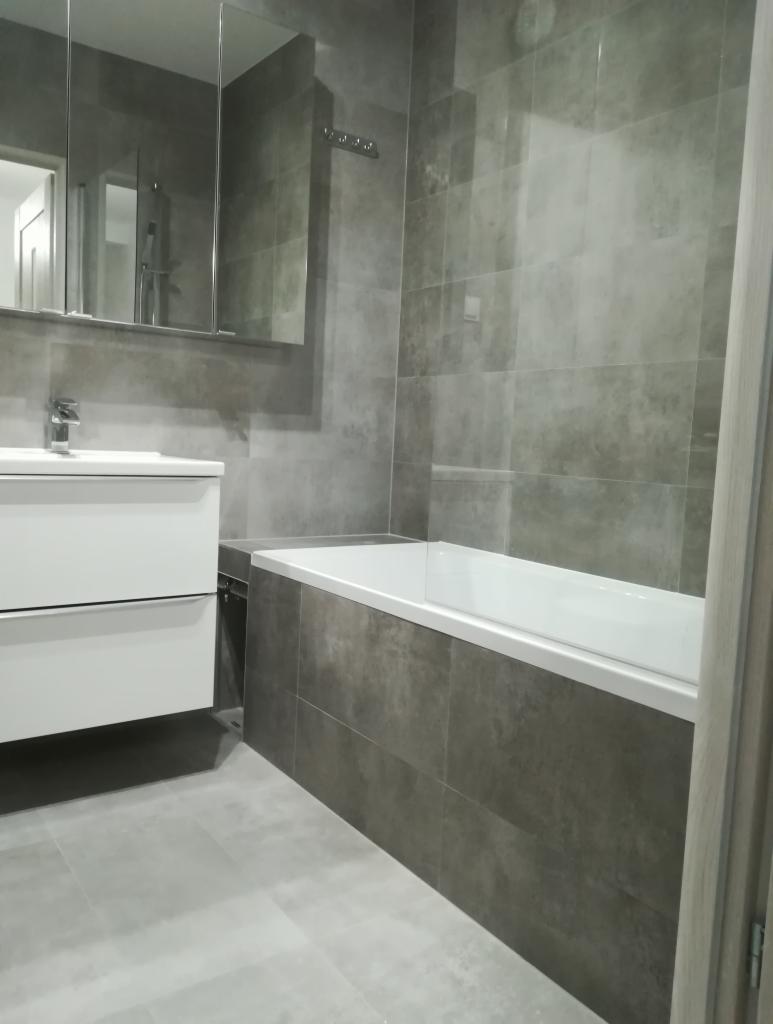 Nowoczesna łazienka w odcieniach szarości z wanną obudowaną płytkami, białą umywalką z szafką i lustrem z szafkami, minimalistyczny design.