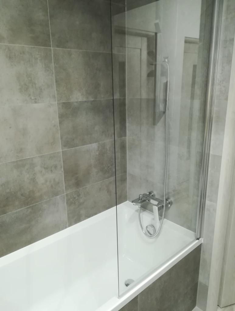 Biała wanna z szarą, teksturowaną okładziną ścienną, chromowaną baterią i przezroczystym, szklanym parawanem nawannowym z widocznym mocowaniem.