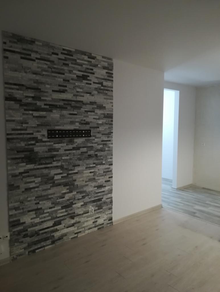 Ściana wyłożona dekoracyjną okładziną kamienną w odcieniach szarości, z zamontowanym uchwytem na telewizor i gniazdkiem elektrycznym, widok na puste wnętrze z panelami podłogowymi.