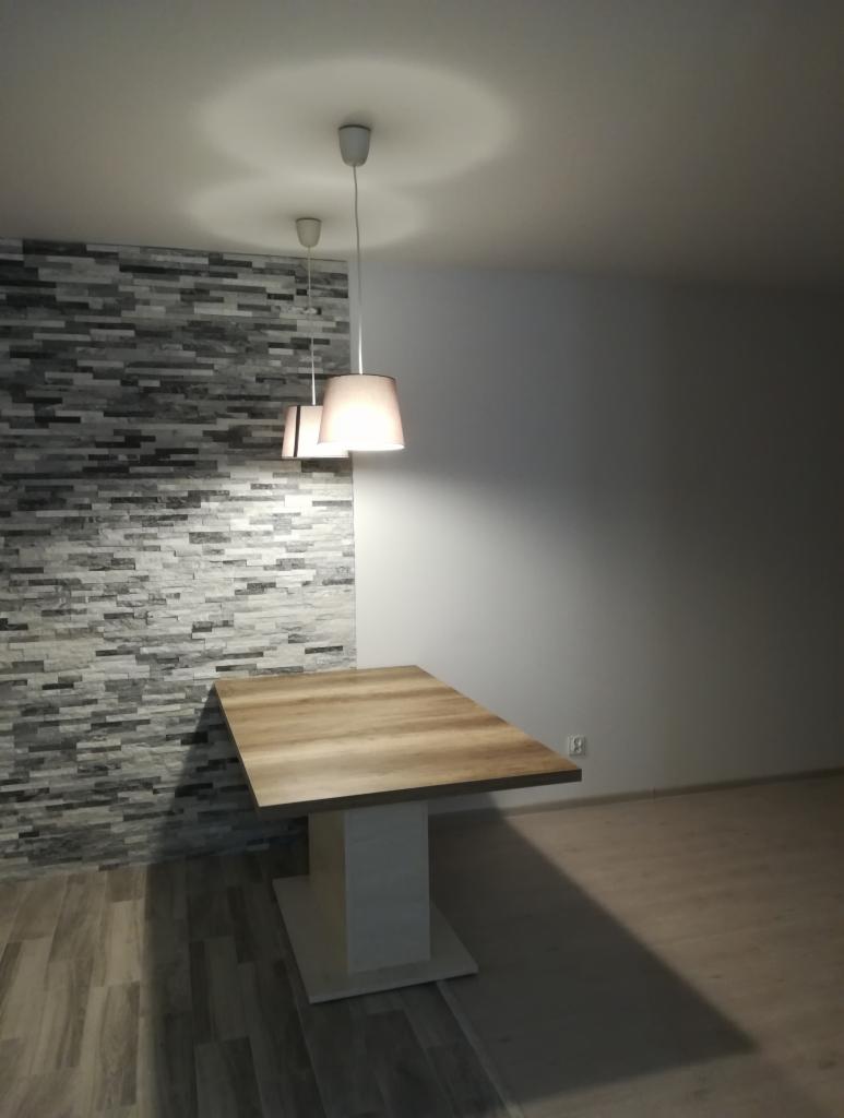 Stół z blatem imitującym drewno, osadzony na białej, kwadratowej nodze, stoi na podłodze wyłożonej panelami, z tłem ściany wykończonej szarymi płytkami imitującymi kamień i białej ściany...