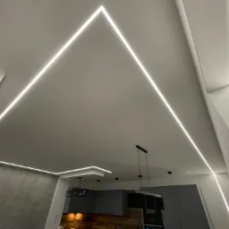 Nowoczesne oświetlenie sufitowe z ledowymi liniami tworzącymi geometryczne wzory, widok na szarą kuchnię z drewnianym blatem i wiszącymi lampami.