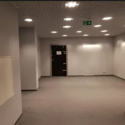 Pomalowane na szaro ściany biura w Chmielniku z widoczną kratownicą sufitową, lampami, drzwiami oklejonymi folią i butelką wody w rogu pomieszczenia.