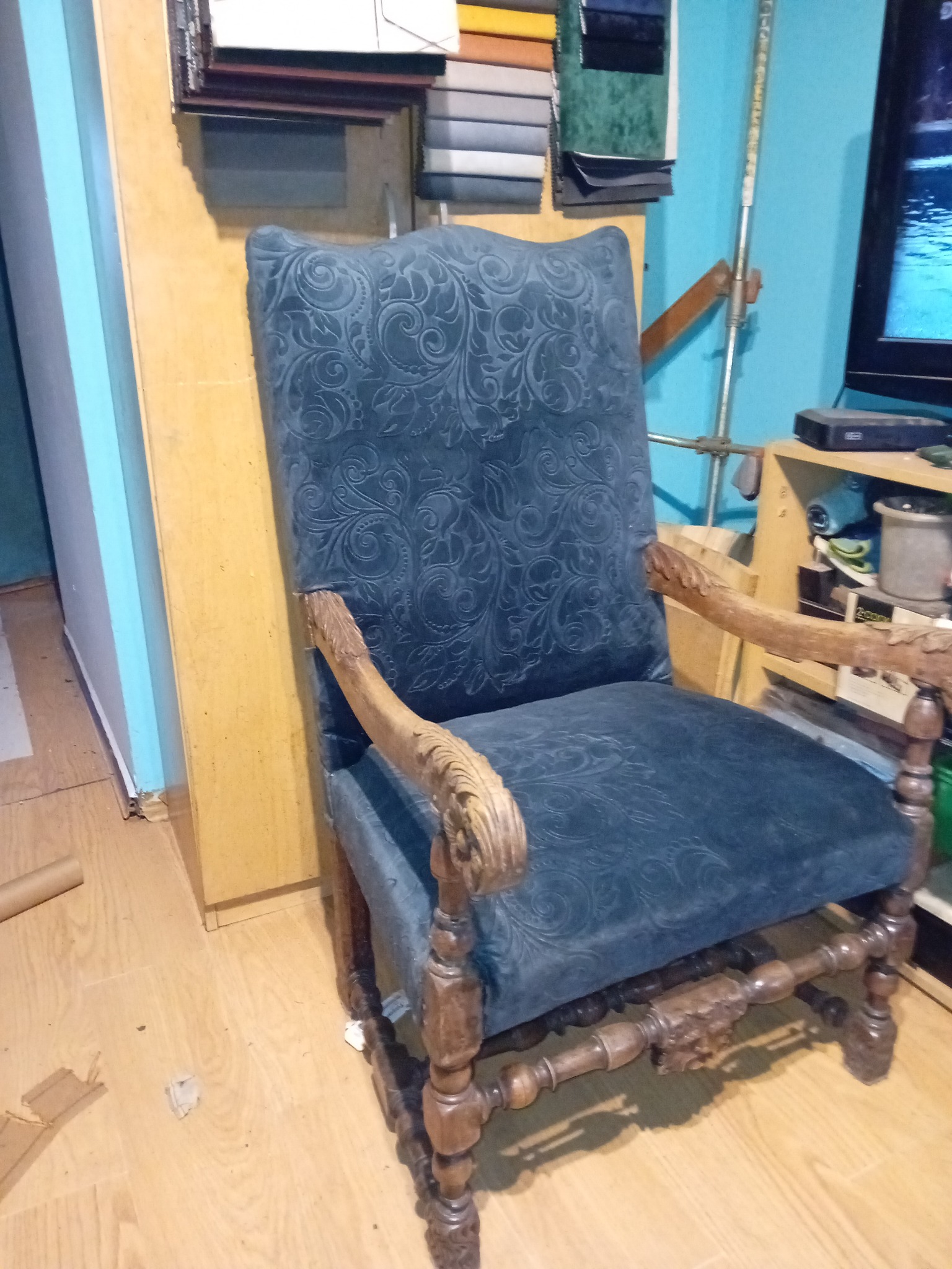 Królewski fotel po renowacji, obity ciemnoniebieskim welurem we wzory, z rzeźbionymi drewnianymi podłokietnikami i nogami, na tle warsztatu.