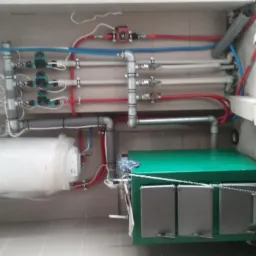 Kotłownia z zielonym kotłem na paliwo stałe, bojlerem owiniętym folią, oraz rozbudowaną instalacją rur miedzianych i plastikowych, widoczne elementy sterujące i zabezpieczające.