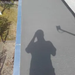 Świeżo zaizolowany, płaski dach pokryty szarą powłoką hydroizolacyjną, z widocznym cieniem osoby wykonującej zdjęcie i obróbkami blacharskimi na krawędzi.