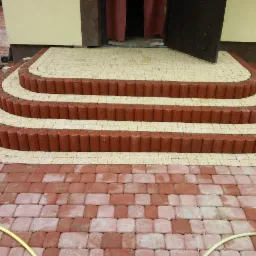 Trzystopniowe schody wejściowe z kostki brukowej w kolorze beżowym i czerwonym, prowadzące do otwartych, ciemnych drzwi wejściowych, z widocznym fragmentem ułożonej kostki brukowej na podjeździe...