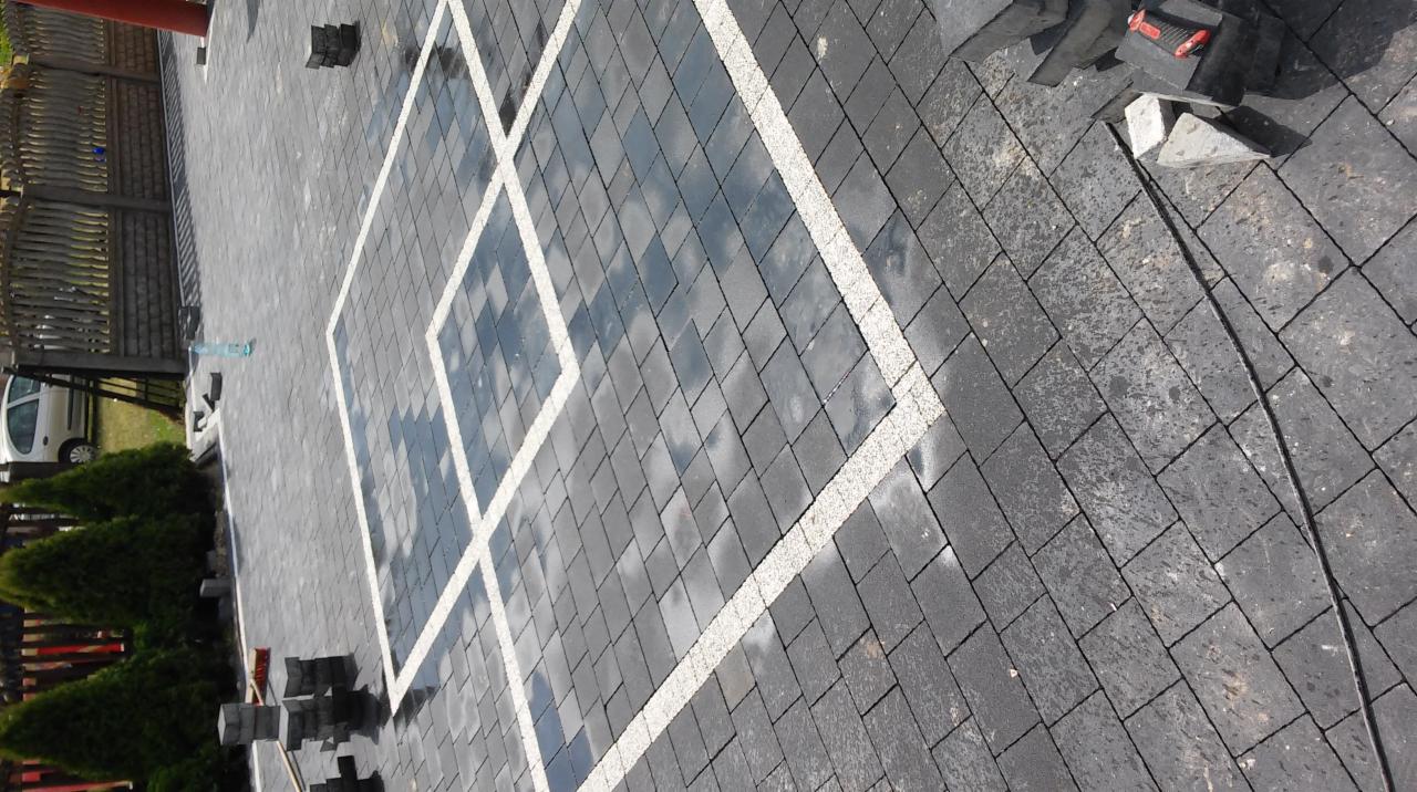 Ułożona kostka brukowa w ciemnoszarym kolorze, z białymi liniami tworzącymi kwadraty, odbijająca niebo. Część kostki jeszcze nieułożona, widoczne narzędzia i butelka wody.