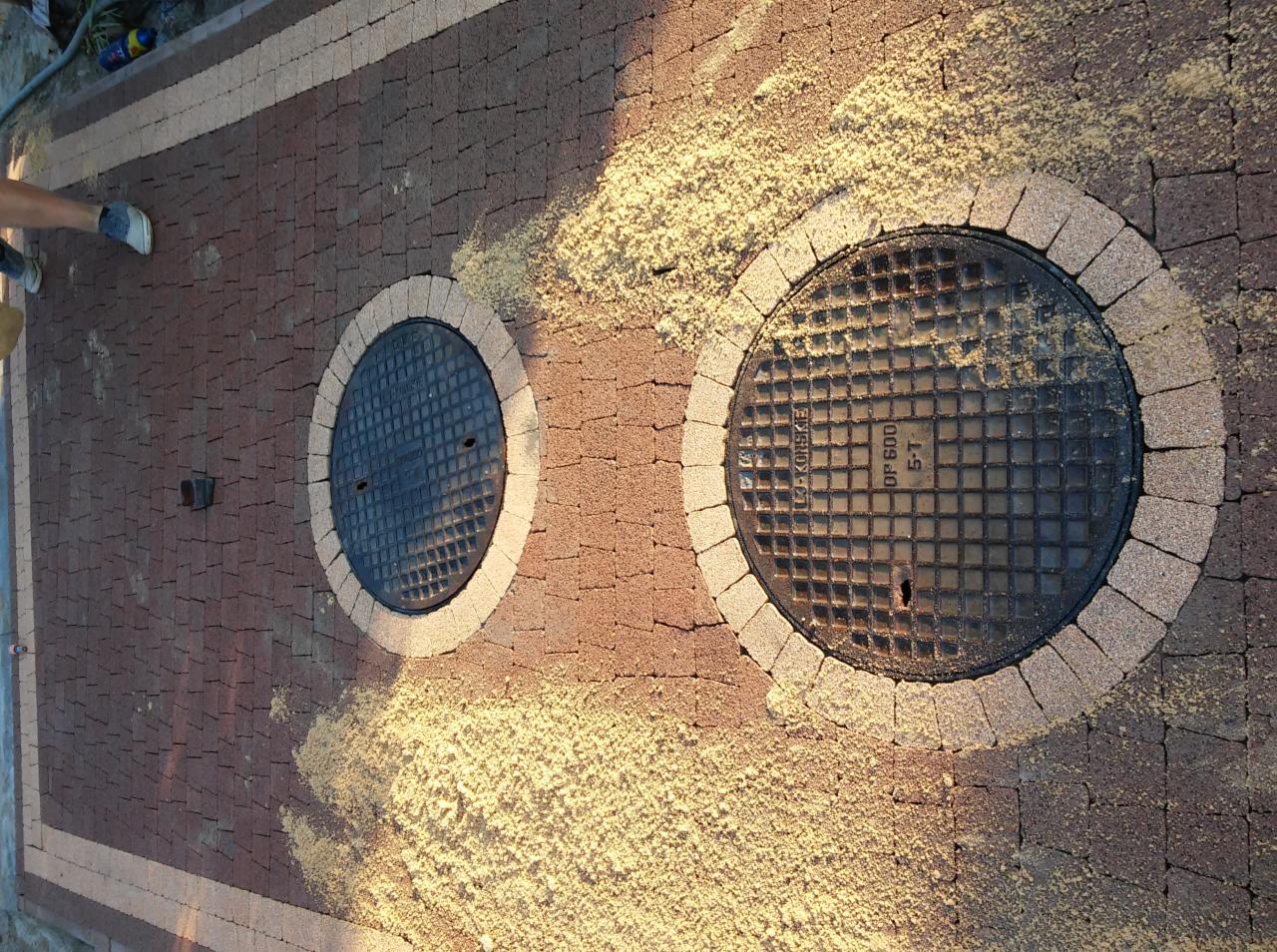 Ułożona kostka brukowa w odcieniach brązu i beżu z dwoma studzienkami kanalizacyjnymi otoczonymi jaśniejszą kostką, zasypana piaskiem.