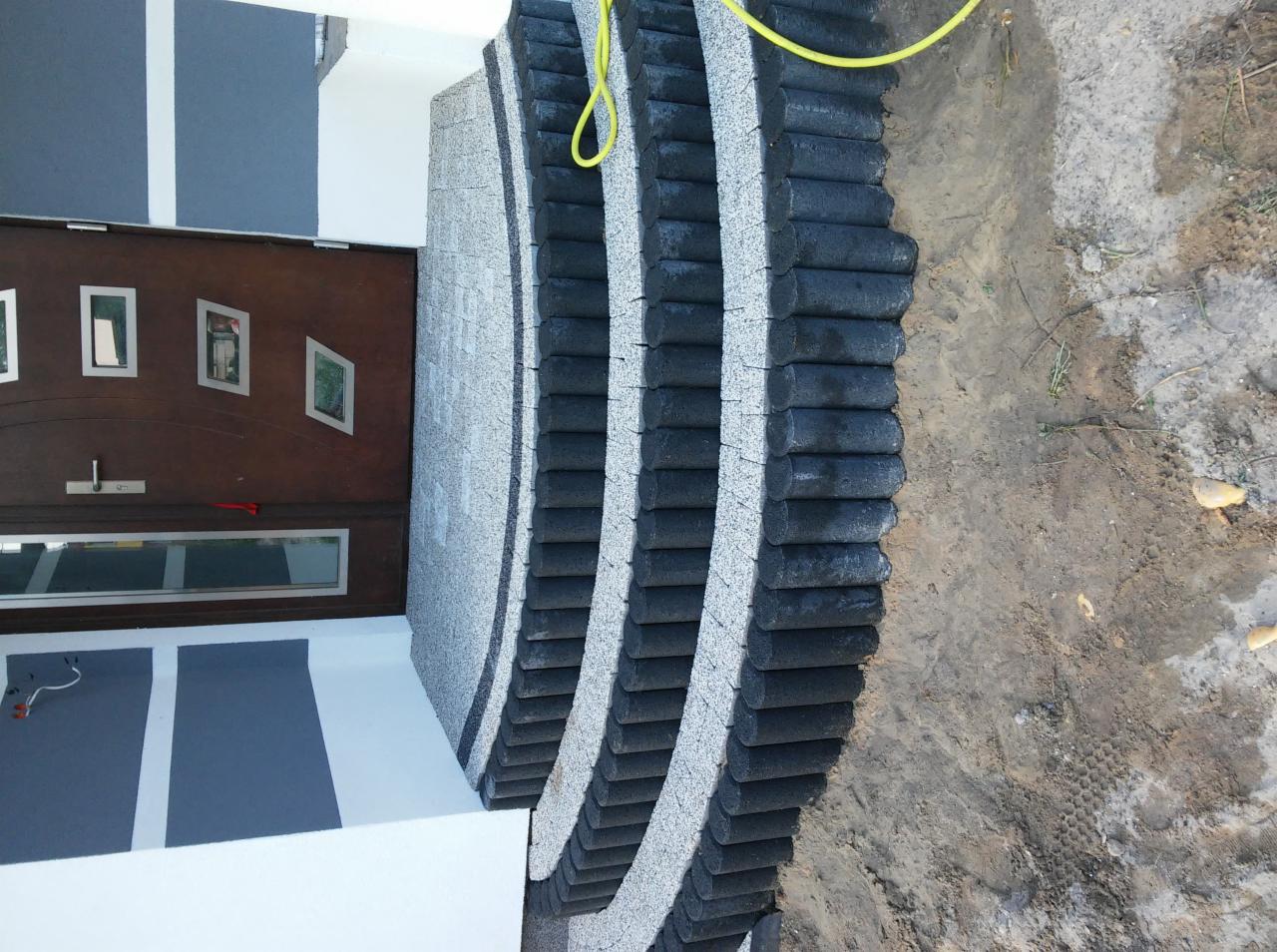 Wykończone schody wejściowe do domu z kostki granitowej i obrzeżami z drewnianych bali, widoczne drzwi wejściowe i fragment elewacji.