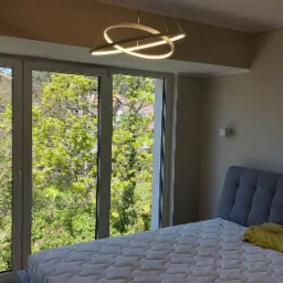 Nowoczesna lampa wisząca LED o geometrycznym kształcie w sypialni z dużym oknem i widokiem na zieleń.