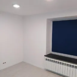 Pomalowane na biało puste pomieszczenie z grzejnikiem pod oknem z granatową roletą i dwiema kwadratowymi lampami sufitowymi.