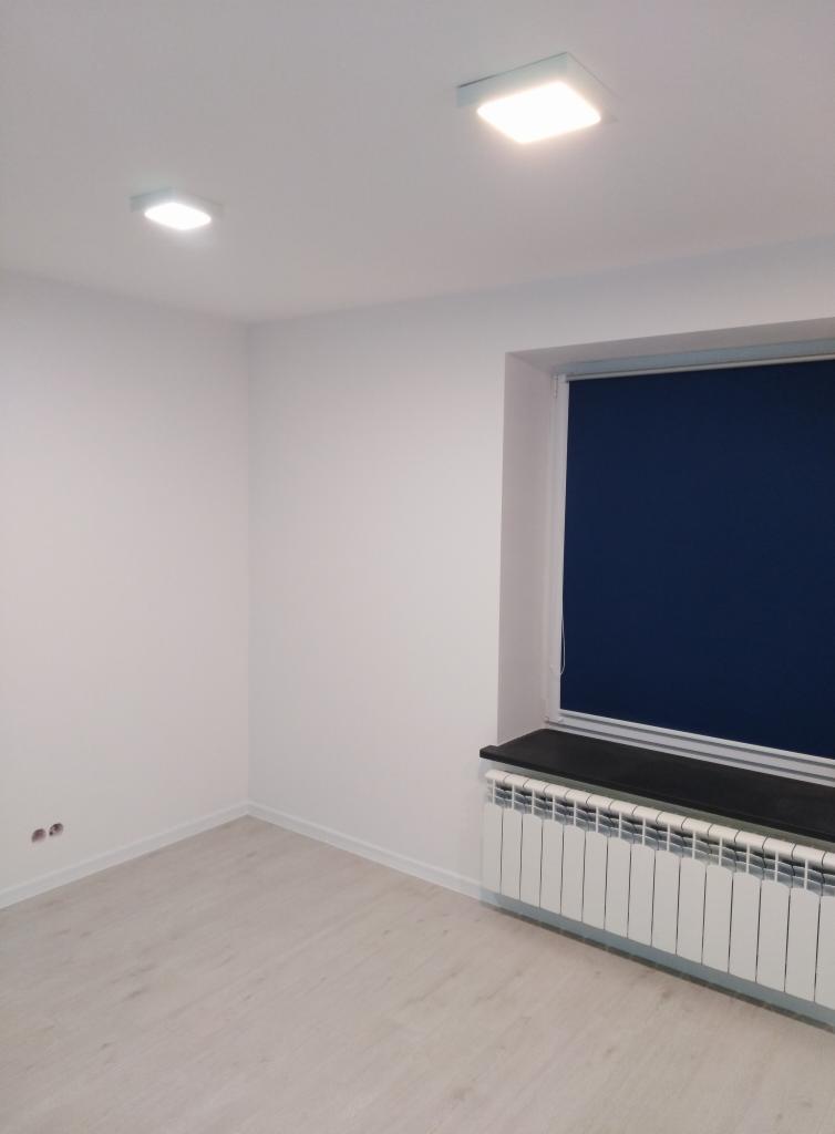 Pomalowane na biało puste pomieszczenie z grzejnikiem pod oknem z granatową roletą i dwiema kwadratowymi lampami sufitowymi.