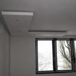 Wykończony sufit podwieszany z gładzią, widoczne kable oświetleniowe, okno z widokiem na parking w zimowej scenerii.