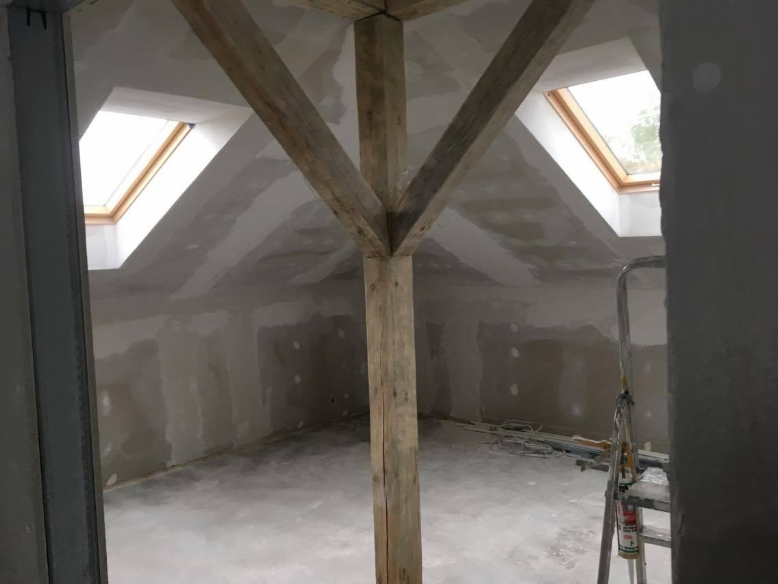 Poddasze w trakcie prac wykończeniowych, widoczne drewniane belki konstrukcyjne, okna dachowe, ściany przygotowane do malowania oraz drabina.