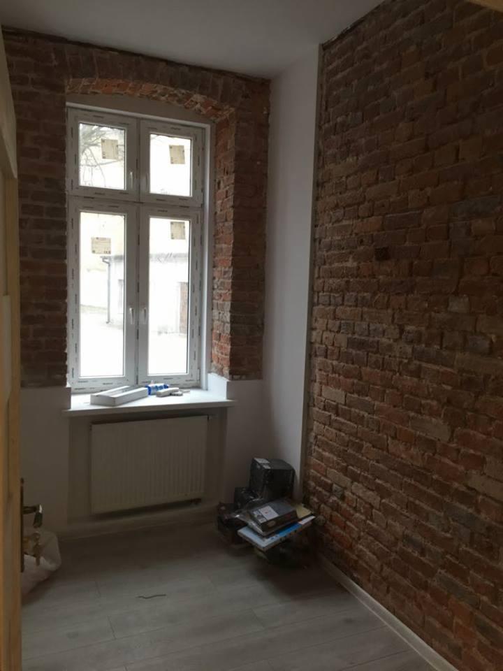 Pokój w trakcie prac wykończeniowych z odsłoniętą ceglaną ścianą i obramowaniem okna, widoczny grzejnik i materiały budowlane na podłodze.