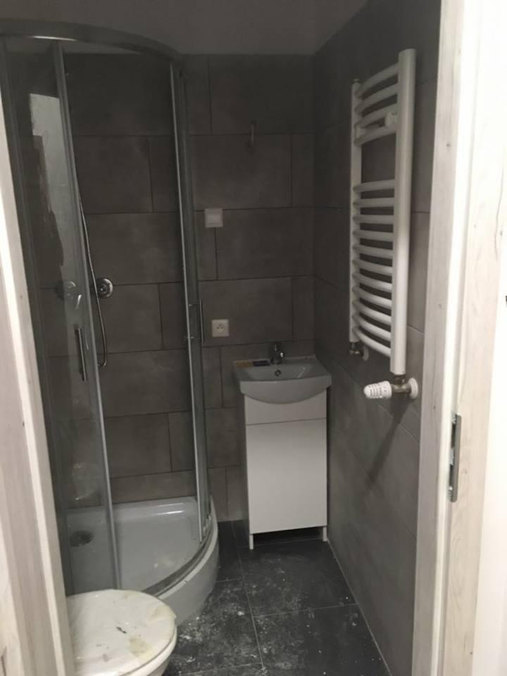 Szara łazienka w trakcie remontu w Łodzi, widoczna kabina prysznicowa, umywalka z szafką i grzejnik drabinkowy, na podłodze widoczne zabrudzenia po remoncie.