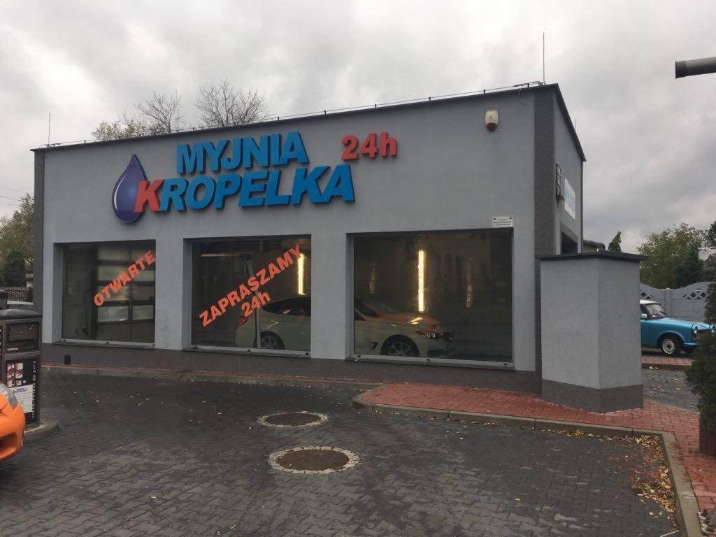 Fasada budynku myjni samochodowej Kropelka z napisem informującym o całodobowej dostępności, widoczne samochody wewnątrz i klasyczny niebieski samochód na zewnątrz