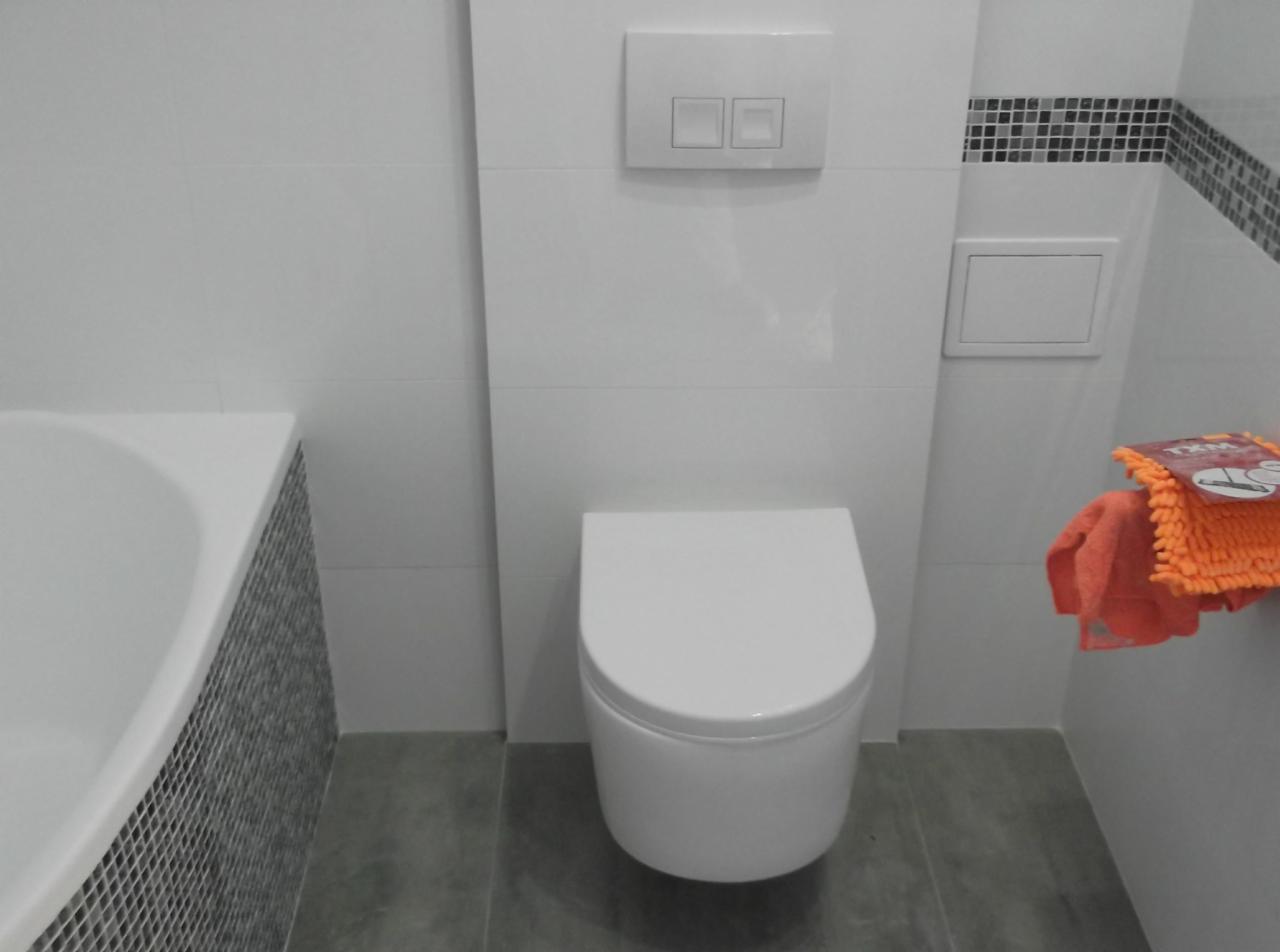 Nowoczesna toaleta podwieszana z białymi płytkami i szarą podłogą, widoczny fragment wanny z mozaiką, pomarańczowy ręcznik po prawej stronie.