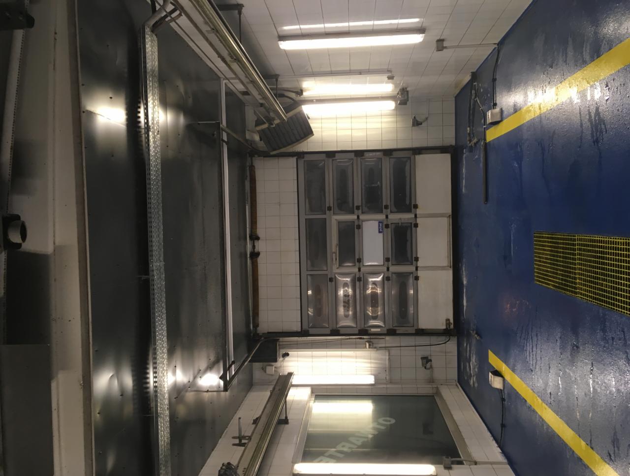 Wnętrze hali z niebieską, mokrą podłogą, białymi kafelkami na ścianach i rzędami metalowych, prostokątnych paneli.