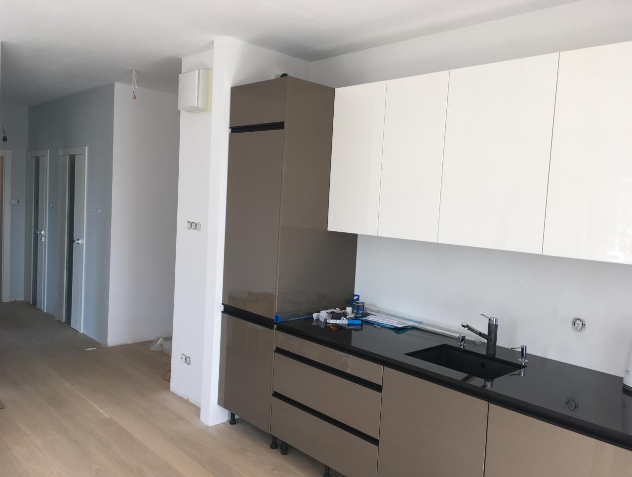 Wnętrze kuchni w trakcie montażu: widoczne szafki dolne w kolorze taupe z czarnym blatem, górne białe szafki wiszące, surowe ściany i podłoga przed wykończeniem, oraz narzędzia na blacie.