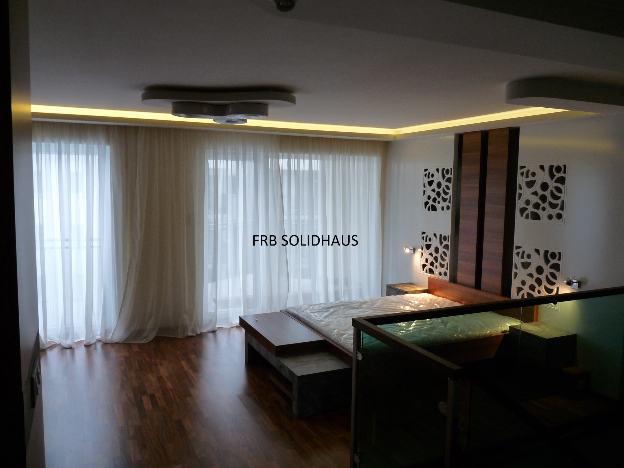 Nowoczesna sypialnia z drewnianą podłogą, dużym oknem z białymi zasłonami, designerskim oświetleniem LED na suficie i dekoracyjną ścianą za łóżkiem.