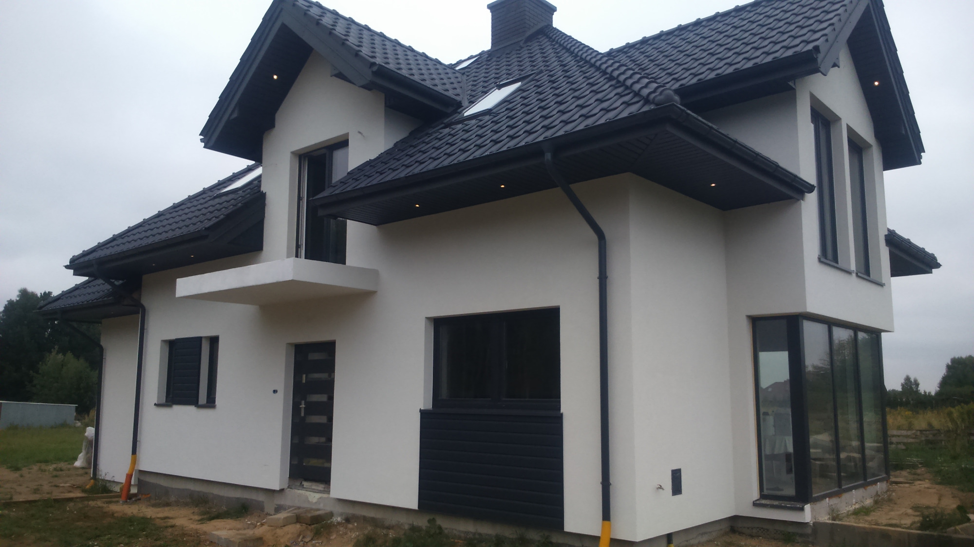 Nowoczesny dom jednorodzinny z białą elewacją i ciemną dachówką, widoczne okna z ciemnymi ramami, oświetlenie zewnętrzne pod dachem, teren wokół domu w trakcie prac wykończeniowych.