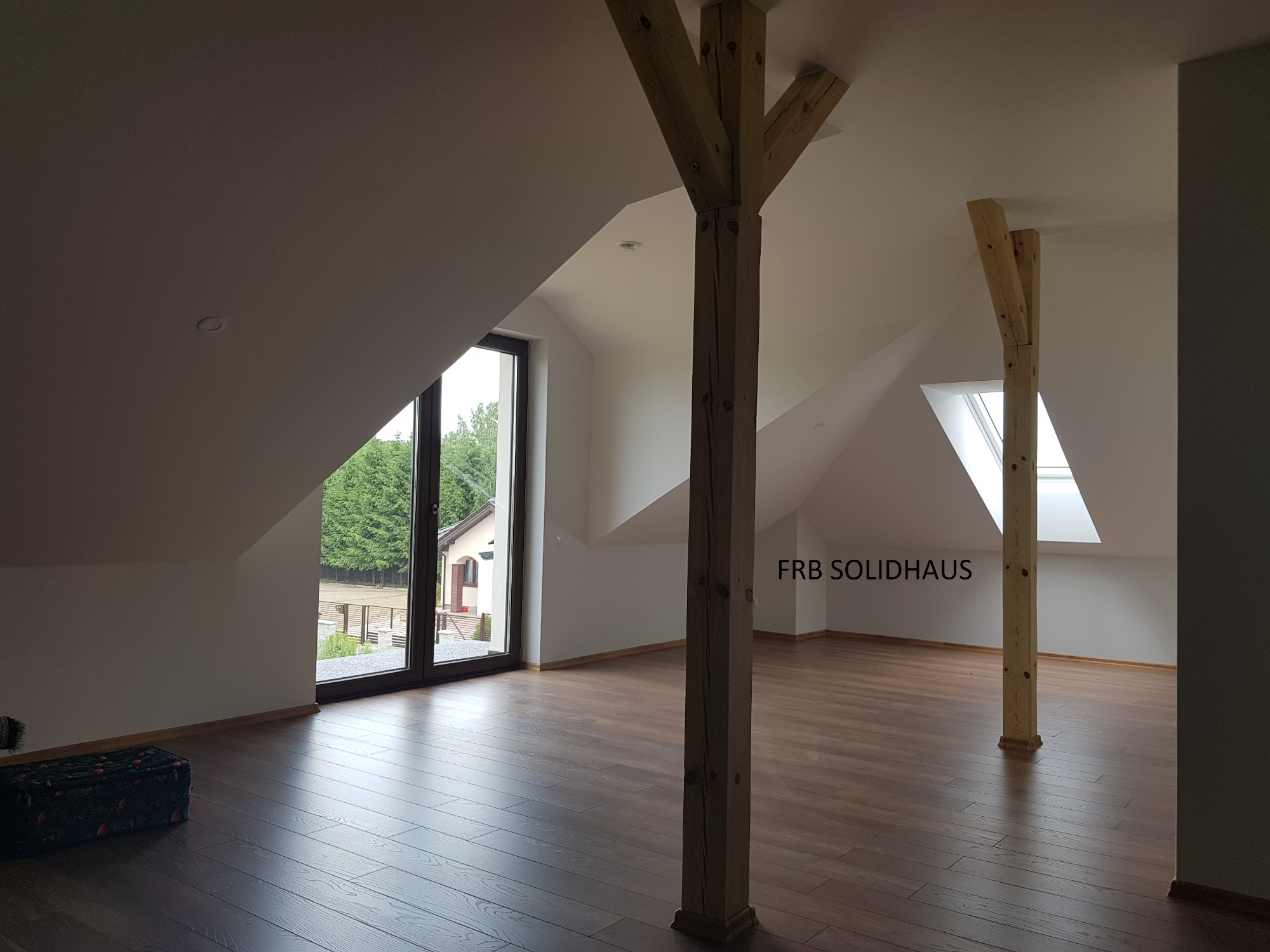 Poddasze po adaptacji z drewnianymi belkami konstrukcyjnymi, widoczne okno dachowe i okno balkonowe z widokiem na ogród, wykończona podłoga z paneli.