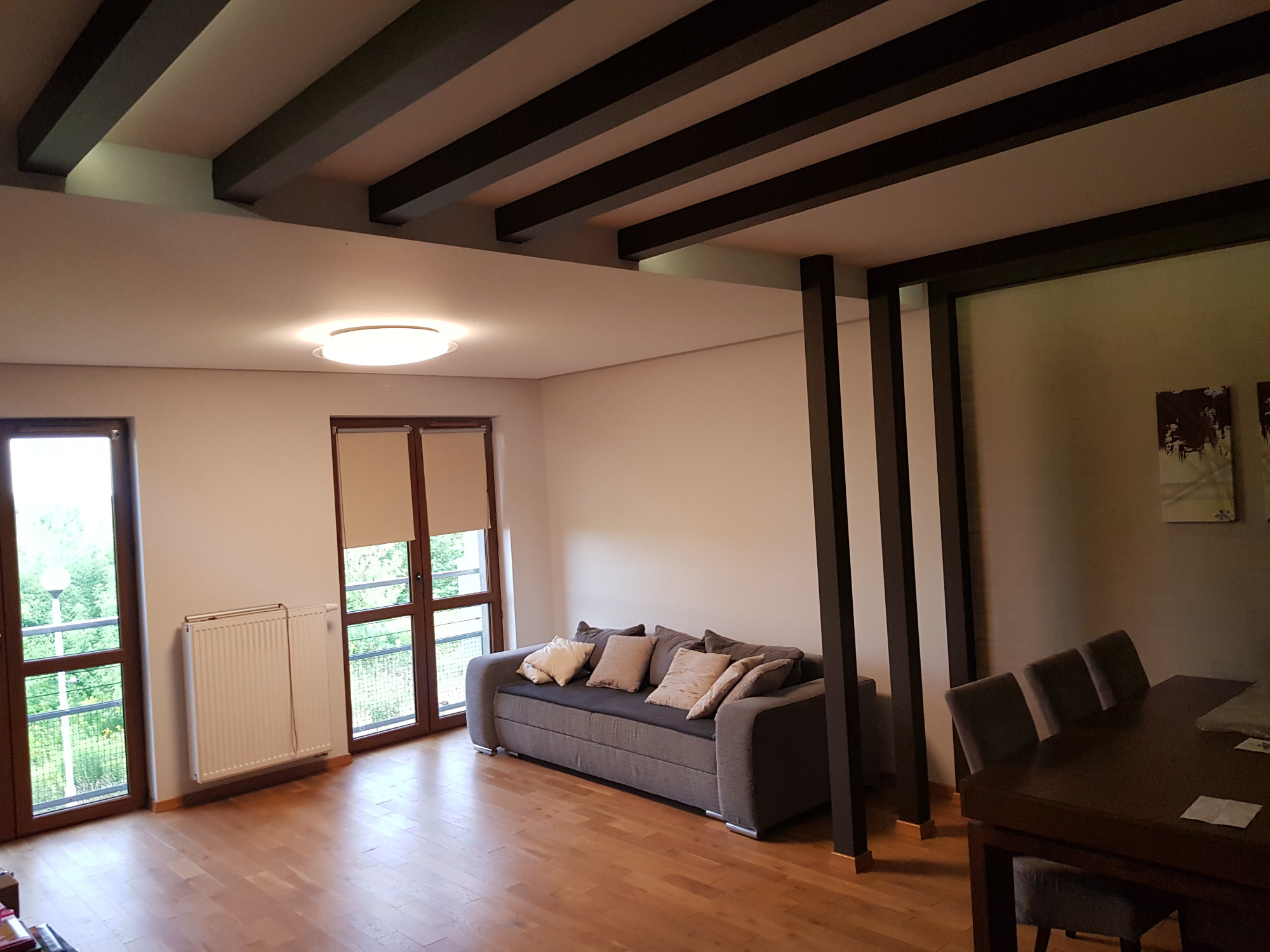 Jasne wnętrze salonu z drewnianą podłogą, szarą sofą z poduszkami, grzejnikiem pod oknem balkonowym i dekoracyjnymi belkami sufitowymi w kolorze ciemnego brązu.
