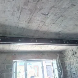 Wzmocnienie stalowym profilem pod stropem w trakcie prac budowlanych, widoczne ślady po deskowaniu na suficie i surowa ściana z cegieł.