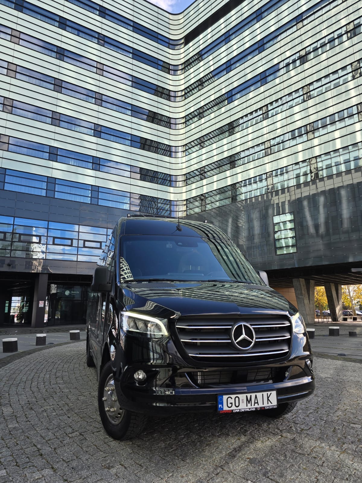 Czarny Mercedes Sprinter z tablicą rejestracyjną GO MAIK zaparkowany przed nowoczesnym budynkiem z licznymi oknami.