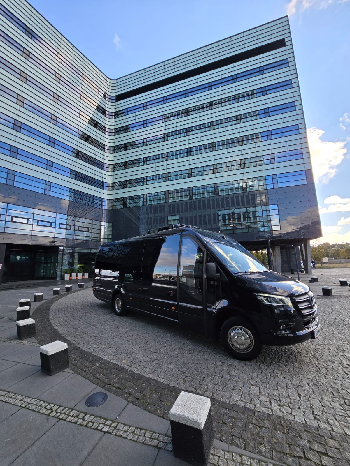 Czarny, luksusowy van Mercedes-Benz z przyciemnianymi szybami, zaparkowany na brukowanym placu przed nowoczesnym biurowcem z licznymi oknami.