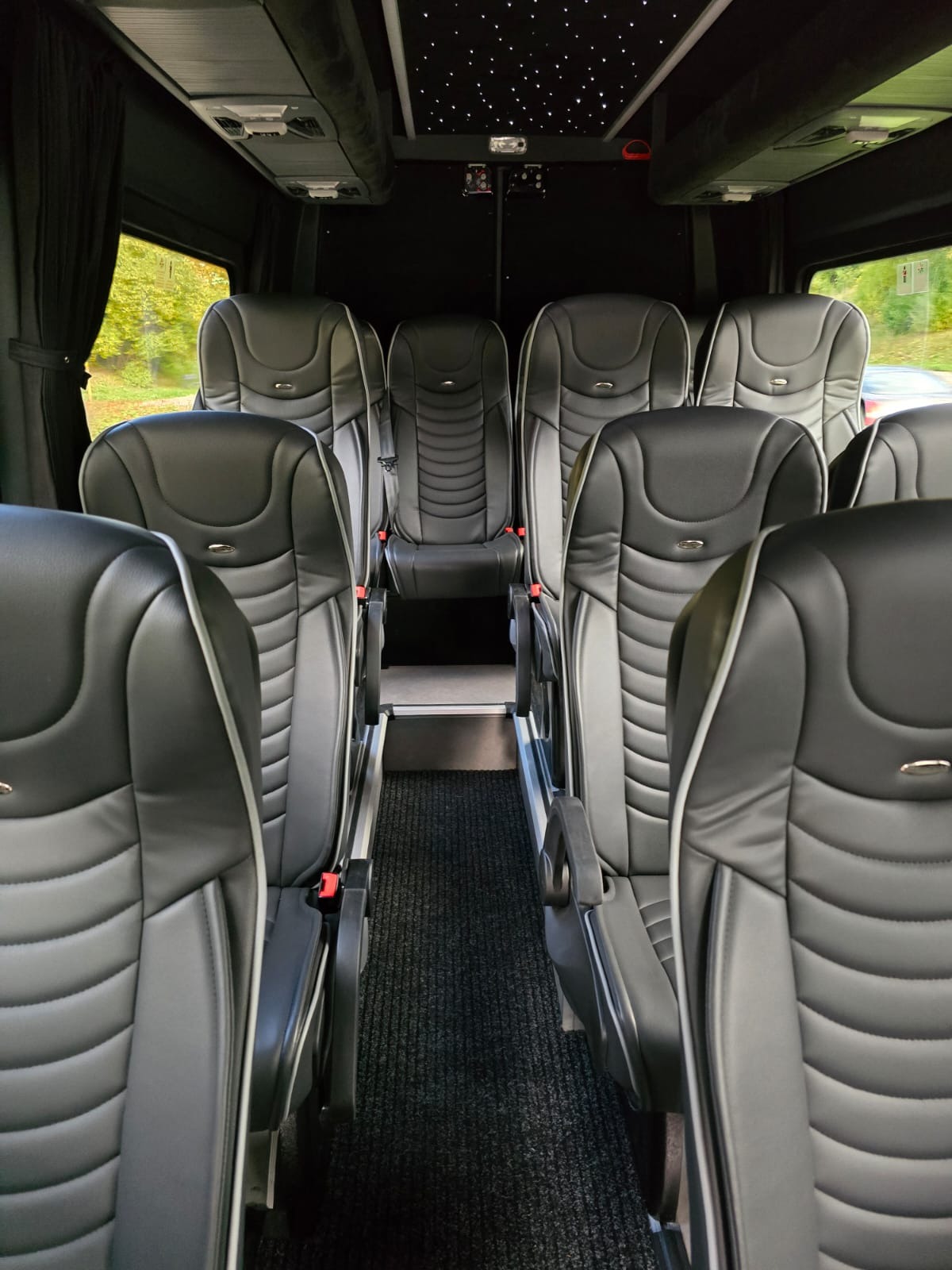 Wnętrze luksusowego busa z czarnymi skórzanymi fotelami z szarymi lamówkami, oświetlenie sufitowe imitujące gwiazdy, widok z tyłu pojazdu.