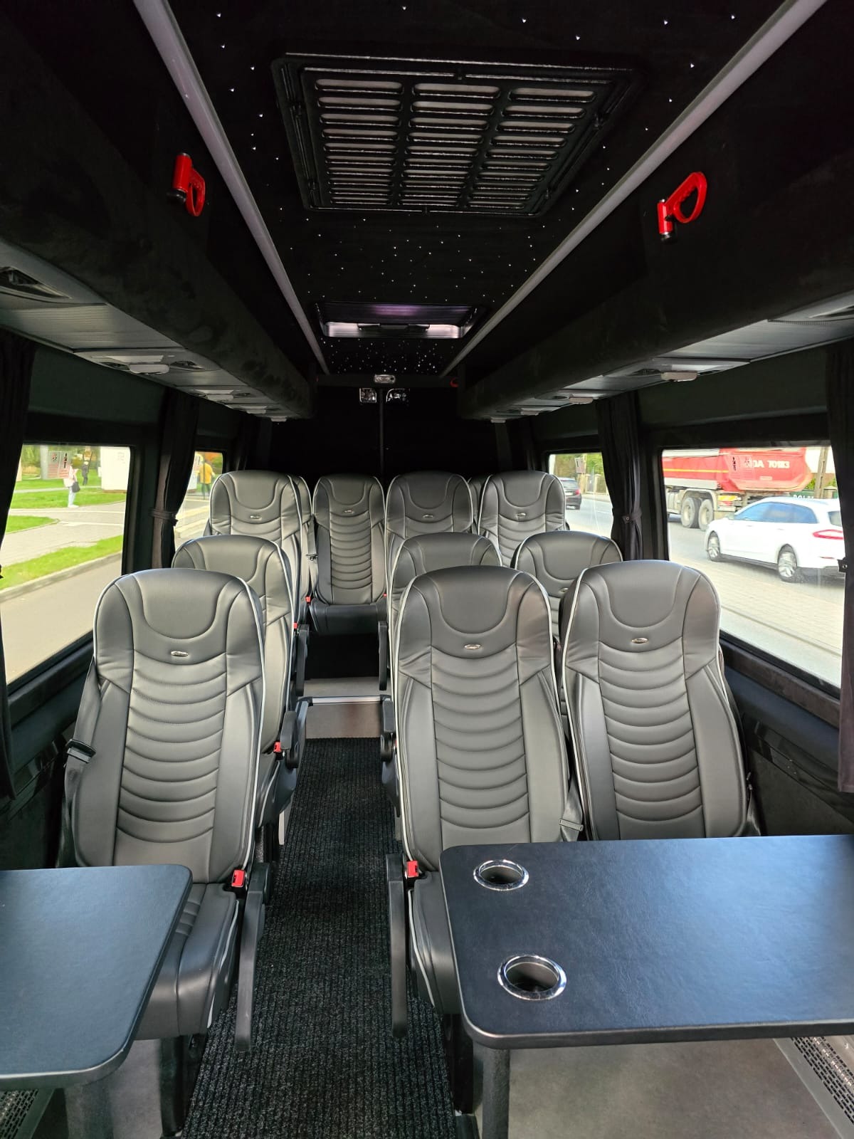 Wnętrze luksusowego busa z czarną tapicerką, skórzanymi fotelami z białymi lamówkami i stolikami z uchwytami na napoje, oświetlenie sufitowe imitujące gwiazdy.