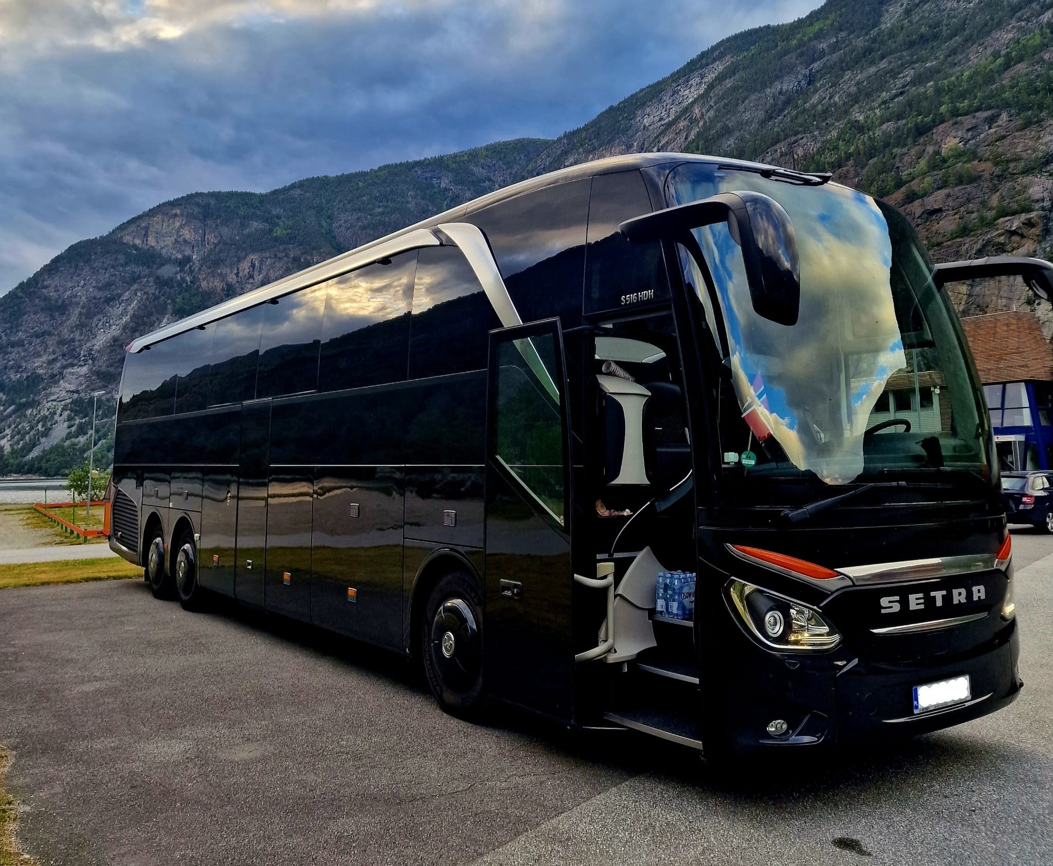 Czarny autokar Setra S 516 HDH z otwartymi drzwiami na tle gór i pochmurnego nieba. Wewnątrz widoczne butelki wody mineralnej.