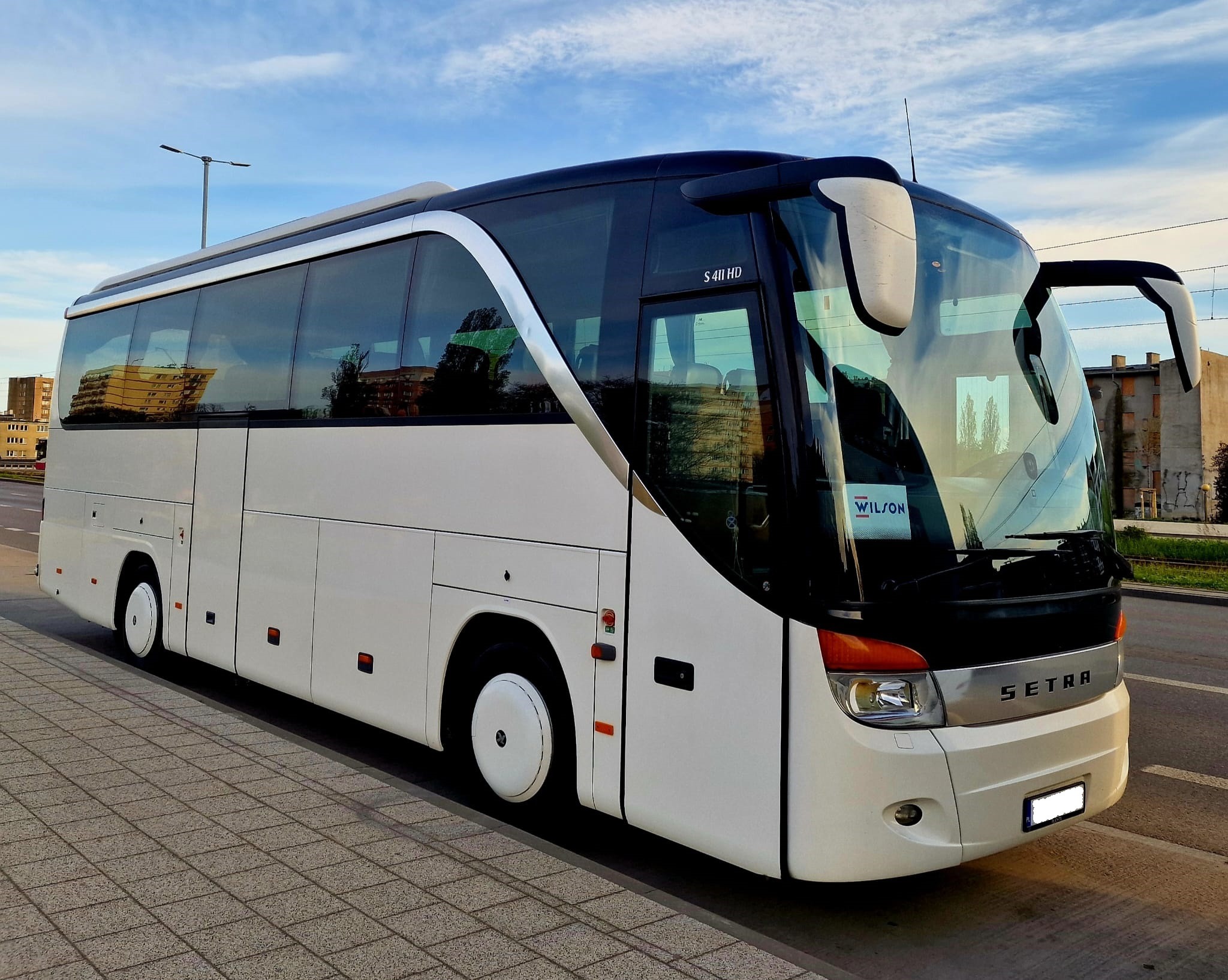 Biały autokar Setra S 411 HD zaparkowany na chodniku z kostki, z widocznym logo firmy Wilson na przedniej szybie, odbijający budynki w oknach.