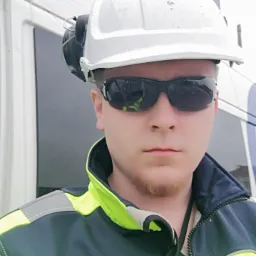 Młody mężczyzna w kasku ochronnym, okularach przeciwsłonecznych i kurtce roboczej z elementami odblaskowymi, pozujący na tle białej ściany z niebieskimi akcentami.