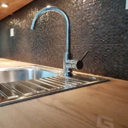 Bateria kuchenna ze stali nierdzewnej zamontowana w zlewozmywaku, na blacie z imitacji drewna, w tle ściana wyłożona czarnymi, heksagonalnymi płytkami ceramicznymi z fakturą.