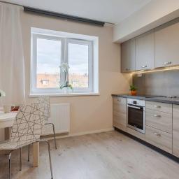 Glazurex - Jasna kuchnia z drewnopodobnymi frontami szafek, szarym blatem i oświetleniem LED pod szafkami wiszącymi, widok na okno z białymi firankami i kwiatami, stół z krzesłami w geometryczne wzory.